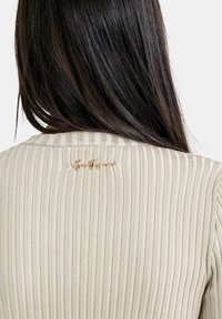 Beige ribbet sweater med rund hals og et gyldent broderet logo på ryggen. Teksturen er blød med et subtilt stribet mønster.