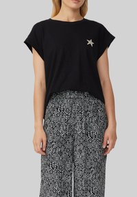 Czarny t-shirt z krótkim rękawem i podwiniętymi mankietami, ozdobiony haftowaną srebrną gwiazdą. W zestawie z wzorzystymi czarno-białymi spodniami typu wide-leg.