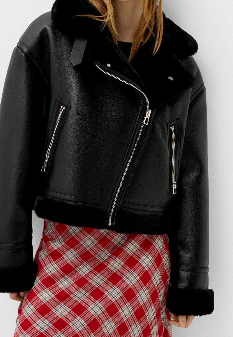 Czarna krótka kurtka ze skóry ekologicznej z kołnierzem z sztucznego futra, ozdobiona srebrnymi zamkami błyskawicznymi z przodu i na kieszeniach. Modelka ma na sobie czerwoną kratowaną spódnicę.