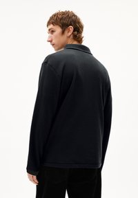 Schwarzes, langärmeliges Polohemd aus strukturiertem Material. Verfügt über einen Kragen und eine lockere Passform. Das Modell ist von hinten zu sehen.