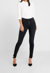 Jeans neri skinny a vita alta, con chiusura frontale a cinque bottoni e tasche. Abbinati a una camicia bianca con colletto e tacchi neri.