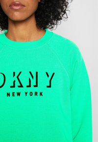 Sudadera verde con cuello redondo y mangas raglán. Presenta una impresión gráfica negra "DKNY NEW YORK" en la parte frontal. Textura suave.