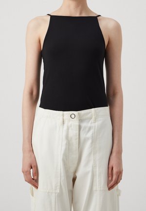 Personne portant un haut noir ajusté sans manches avec des bretelles spaghetti et un pantalon blanc ample à taille haute avec des poches devant.