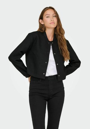 Jeune femme aux longs cheveux bruns portant une veste noire à boutons, une chemise blanche et un pantalon noir taille haute, se tenant les mains sur les hanches.