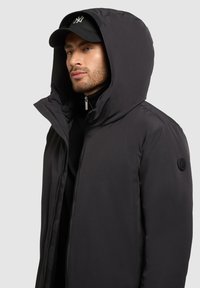 Fekete puffdzseki magas gallérral és nagy kapucnival, elöl cipzárral és a bal ujján logóval.