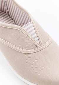 Zapato de lona beige sin cordones con un upper texturizado, que cuenta con un panel elástico en forma de V y un forro interior a rayas. Suela de goma blanca.