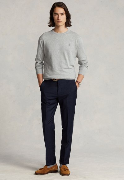 Giovane uomo in piedi con le mani in tasca, indossa una camicia grigio chiaro a maniche lunghe, pantaloni scuri, mocassini marroni e un orologio da polso.