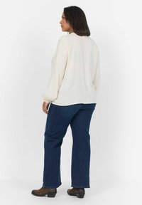 Blusa crema con maniche lunghe a sbuffo, abbinata a jeans blu scuro a gamba dritta e stivali alla caviglia marroni, vista da dietro.
