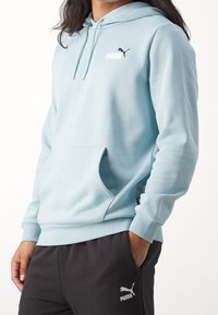 Vaaleansininen huppari, jossa on etutasku, kiristysnauhallinen huppu ja pieni musta Puma-logo rinnassa. Pehmeää kangasta ja resorit hihansuissa.