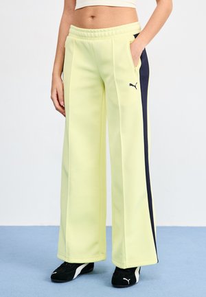 Puma wide-leg sweatpants in pale yellow, featuring side pockets and navy accents. Soft fabric with a relaxed fit. Black footwear completes the look.