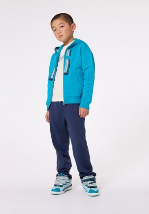 Tyrkise jakke med glidelås og teksturerte striper, hvit t-skjorte, marineblå joggebukser og matchende sneakers med fletter i snøringen.