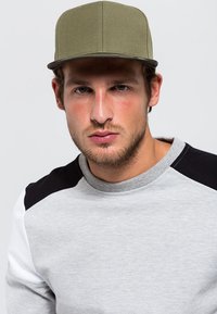 Cappello verde oliva con visiera piatta, realizzato in un tessuto testurizzato, abbinato a una felpa grigia con blocchi di colore bianco e nero sulle maniche.