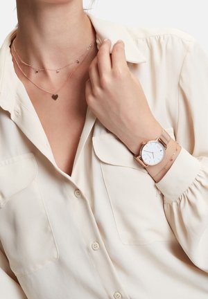 Femme portant plusieurs colliers en forme de cœur en or, une chemise beige boutonnée et une montre à bracelet en mesh en or rose à son poignet gauche.