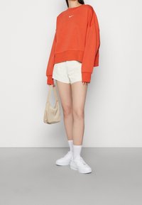 Pomarańczowa krótka bluza z białym logo, zestawiona z białymi szortami, białymi trampkami i jasnobeżową torebką na ramię.