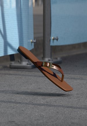 Barna bőr flip-flop arany csattal szürke járda felett lebeg, fémrudak és kék kerítés közelében.