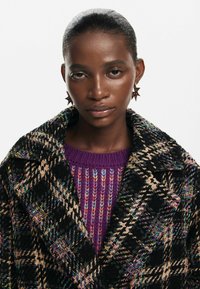Black and tan plaid coat with a soft texture, layered over a purple knitted sweater. Star-shaped earrings add a distinctive accent.