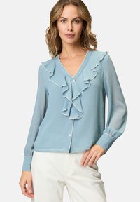 Femme portant un blouse bleu clair avec un col en V à volants et de longues manches transparentes, assortie à un pantalon blanc cassé sur un fond uni.