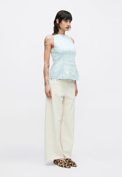 Γυναίκα με τατουάζ που φοράει αμάνικη ανοιχτομπλε μπλούζα, λευκό παντελόνι φαρδιάς γραμμής και παπούτσια τύπου slip-on με λεοπάρ σχέδιο, στέκεται μπροστά σε απλό φόντο.