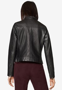 Musta nahkatakki, jossa on istuva muotoilu, hillitty kiilto, vetoketjillä varustetut hihat ja lyhennetty helma. Silkkinen pinta minimaalista yksityiskohtaa.