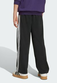 Pantalons de survêtement noirs en tissu doux avec des rayures blanches sur les côtés. Dotés d'une coupe décontractée et d'une texture lisse, portés avec des baskets blanches.
