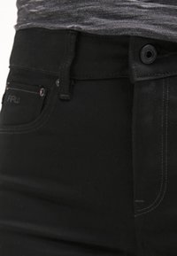 Jean en denim noir avec une coupe slim, des détails de poches avant et des surpiqûres contrastantes le long des coutures. Fermeture par bouton et zip.