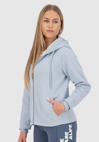 Felpa con zip di colore azzurro, morbida al tatto, con cappuccio regolabile, tasche laterali e polsini a costine. Indossata con pantaloni blu abbinati con testo.