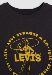 Černé bavlněné tričko s kulatým výstřihem, se světlým žlutým grafickým motivem kovboje a textem "Levi's" a "Levi Strauss & Co." v kruhovém vzoru.