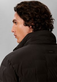 Jeune homme aux cheveux bouclés et foncés vu de profil, portant une veste noire Calvin Klein avec le col relevé.