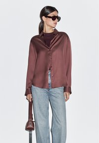 Long-sleeve, burgundy satin shirt with a button-down front, paired with light blue denim jeans and a dark handbag. Sunglasses accentuate the look.