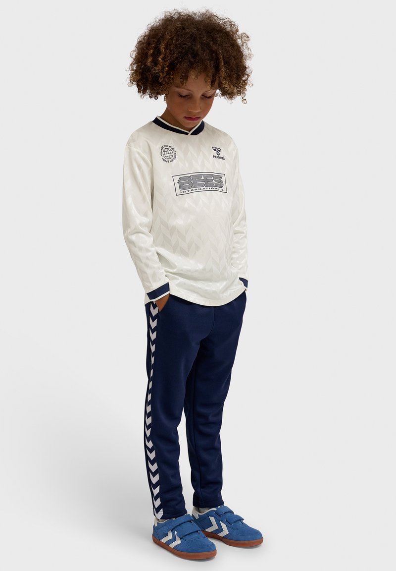 Enfant aux cheveux bouclés portant un maillot de sport blanc à manches longues et un pantalon de survêtement bleu marine, debout les mains dans les poches en regardant vers le bas.
