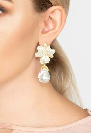 Gold flower-shaped earring with small crystals and a large dangling baroque pearl worn on a woman's left ear.