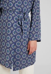 Blå mönstrad morgonrock med cirkulära mönster i turkos och vitt, med knytbar midja, långa ärmar och en mjuk textur.