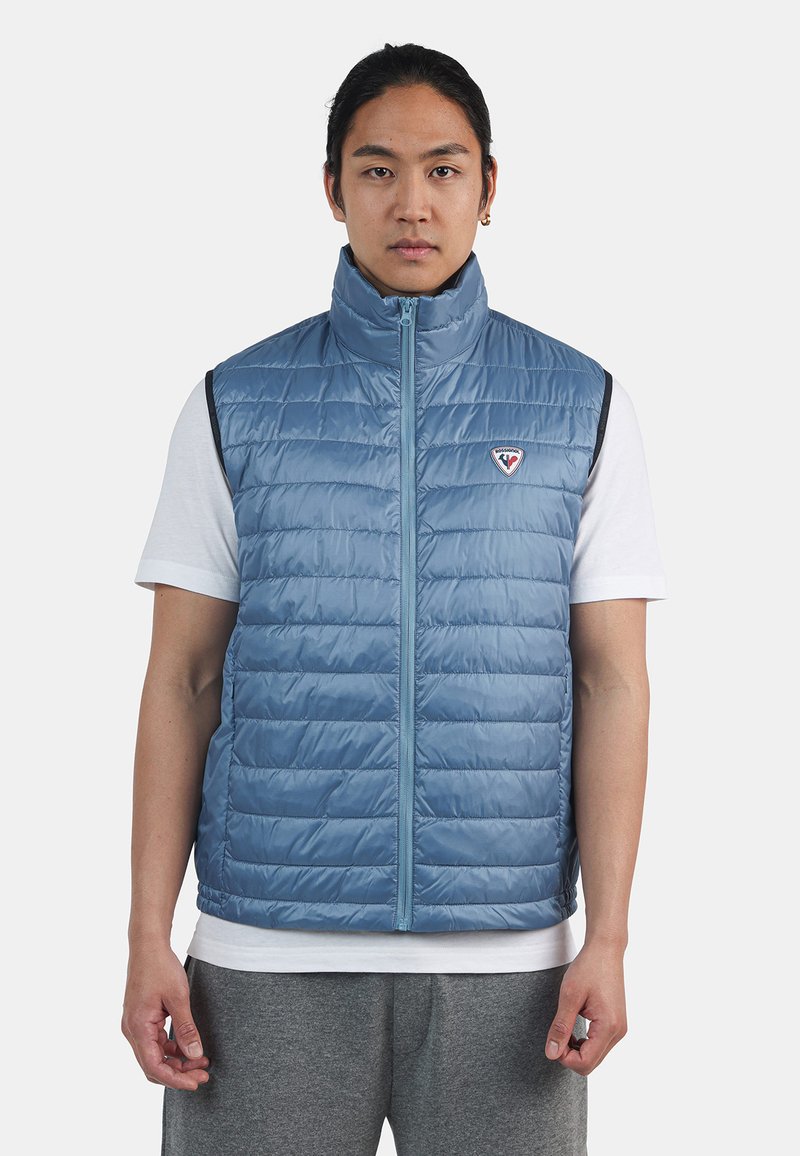 Ljusblå quiltad väst med hög krage, framdragkedja och logopatch. Har vadderade sektioner och kontrasterande mörk armhålskant.