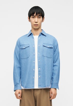 Homme portant une chemise en denim bleu clair boutonnée sur un haut blanc et un pantalon marron, debout devant un fond blanc uni.