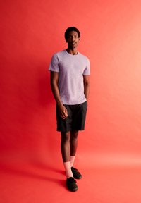 Paarse heide crew-neck t-shirt, zwarte shorts en zwarte sneakers, aangevuld met lichtroze sokken, tegen een rode achtergrond.