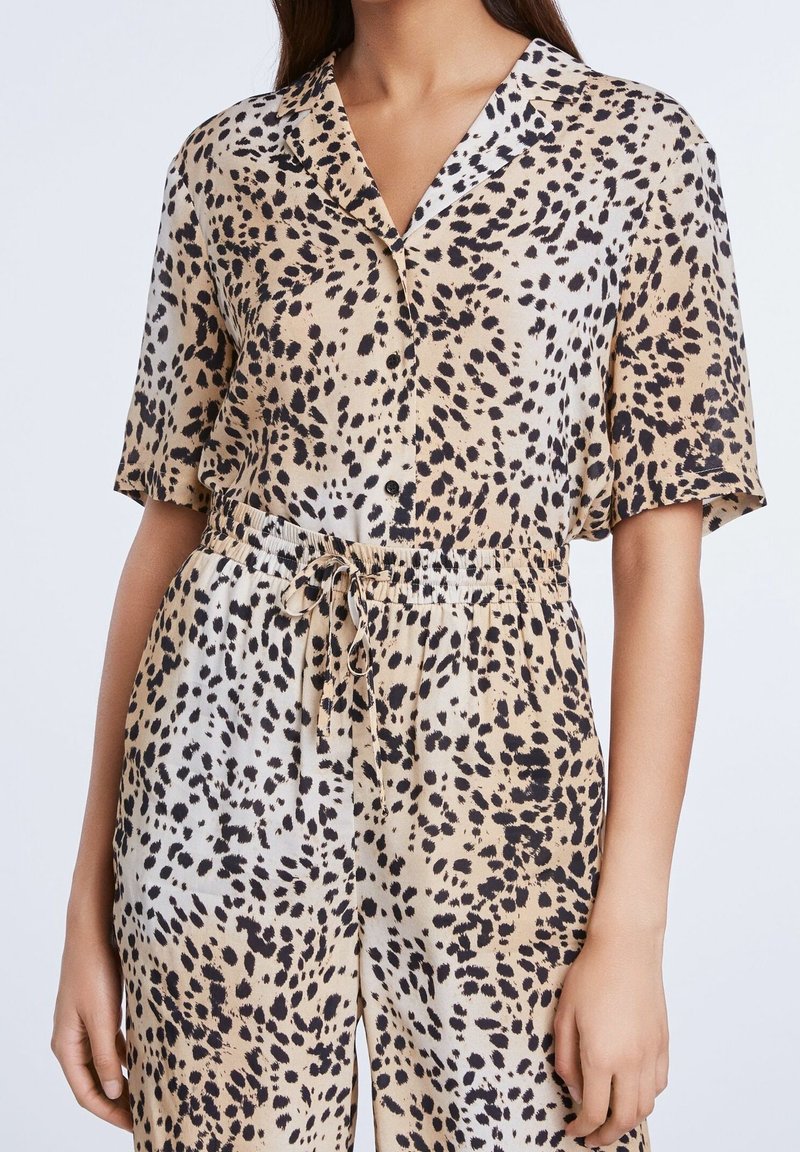 Combinaison à manches courtes en tissu beige présentant un motif tacheté noir et marron, avec un col cranté et un détail taille à cordon de serrage.