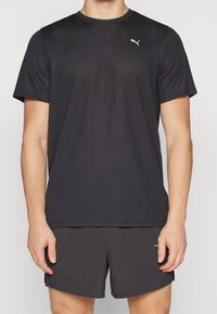 Man i svart Puma t-shirt med kort ärm och matchande svarta shorts, stående framför en enkel bakgrund.