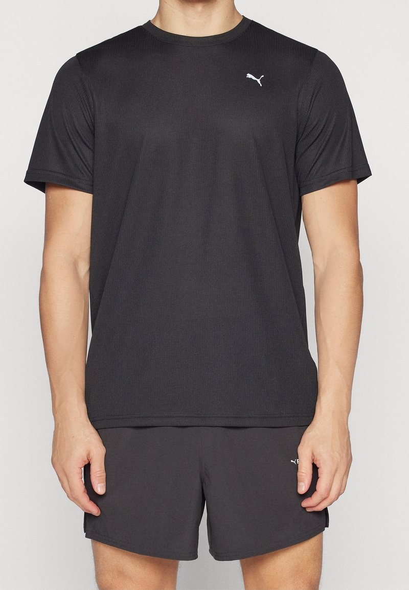 Man i svart Puma t-shirt med kort ärm och matchande svarta shorts, stående framför en enkel bakgrund.