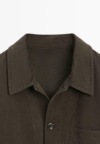 Bruin geknoopt overhemd met een knoopsluiting, borstzak en textuurstof. De kraag heeft een lichte rol voor een gestructureerde uitstraling.