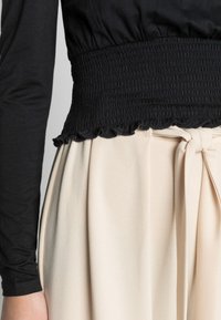 Top preto de mangas compridas com uma cintura franzida, combinado com uma saia plissada bege com um cinto atado. Textura de tecido suave e fluida.