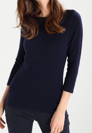 Kobieta ubrana w dopasowaną granatową bluzkę z długim rękawem do łokcia oraz ciemne spodnie w prążki na białym tle.