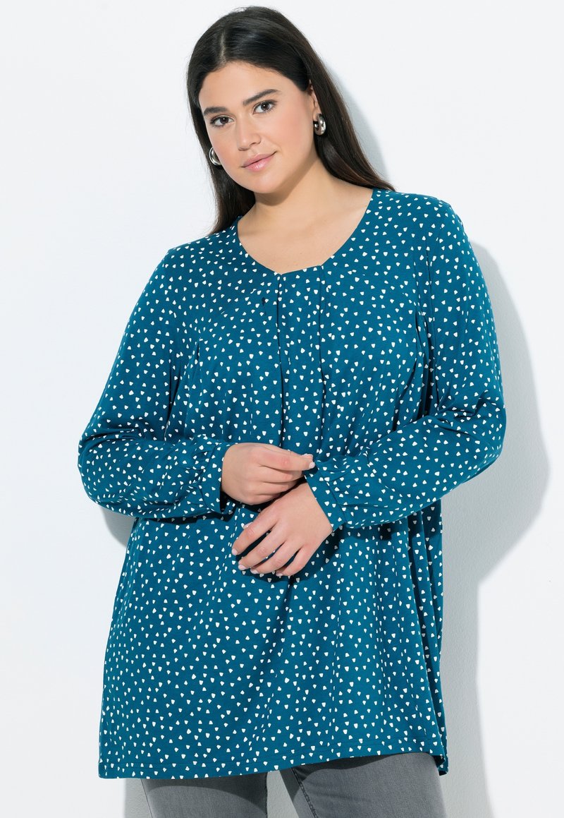 Jonge vrouw met lang donker haar, gekleed in een turquoise blouse met witte abstracte stippen en een grijze broek, staand tegen een witte achtergrond.