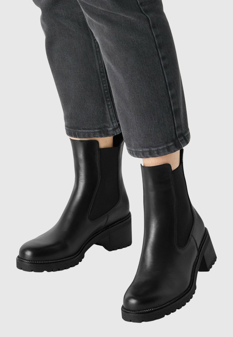 Bottes noires en cuir avec talons épais portées avec un jean court gris foncé sur fond uni.
