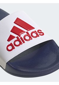 Sandalia deslizante blanca con una correa texturizada que presenta un logotipo de "adidas" rojo en relieve. Plantilla texturizada de color azul oscuro y suela lisa.