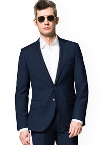 Granatowa marynarka w dopasowanym kroju, z dwoma przednimi kieszeniami i pojedynczym zapięciem na guzik, w połączeniu z białą koszulą zapinaną na guziki oraz okularami przeciwsłonecznymi.