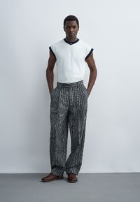 Homme debout, les mains dans les poches, portant un haut blanc sans manches avec bordure marine, pantalon large rayé noir et blanc, et chaussures marron.
