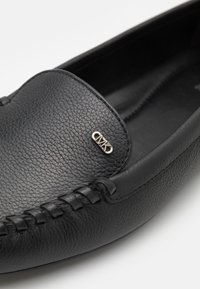 Mokassinschuhe aus schwarzem Leder mit strukturiertem Obermaterial, gesteppten Details und einem kleinen metallischen Logoakzent auf der Vorderseite.