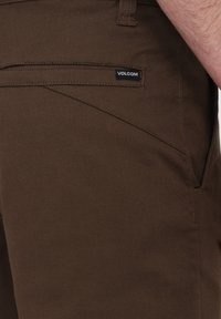 Brune bukser lavet af holdbart stof med en glat tekstur. Har et elegant design og en sidelomme med et sort "Volcom"-mærke.