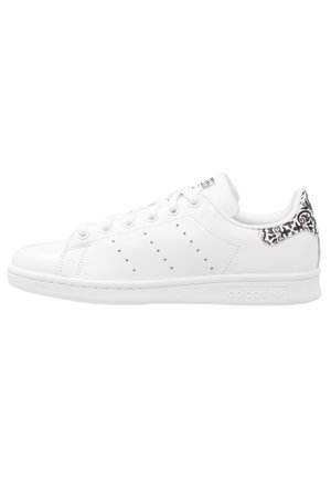 Hvid Adidas lav-top sneaker med perforerede tre striber og sort blomsterprint på hælkappen, set fra siden.