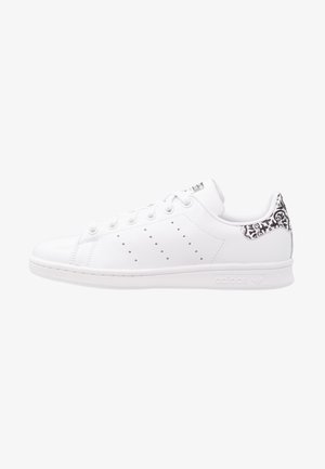 Biała niska biała tenisówka Adidas z perforowanymi trzema paskami i czarnym kwiatowym wzorem na zapiętku, widziana z boku.